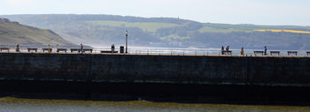 This landscape photograph captures the Whitby Breakwater promenade in Yorkshire during a spring afternoon. The promenade is the main subject of the image, with several people walking or cycling along its length, enjoying the fresh air and scenic views. The stone breakwater is bordered by benches and lamps, which add character to the promenade. In the background, rolling hills with lush greenery and farmland typify the Yorkshire countryside, with coastal cliffs descending towards the sea. The lighting and shadows in the image reflect the afternoon sun, giving the photograph a vibrant yet tranquil atmosphere.